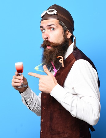 handsome bearded pilot or aviator man with long beard and mustache on funny face holding glass of alcoholic shot in vintage suede leather waistcoat with hat and glasses on blue studio backgroundの写真素材