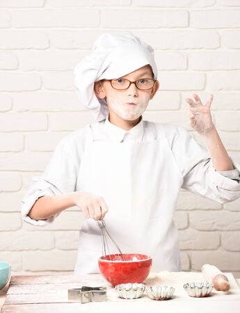 young boy small cute cook chef in white uniform and hat on happy face with glasses standing near table with rolling pin molds for cakes red bowl and playing with flour on brick wall backgroundの写真素材