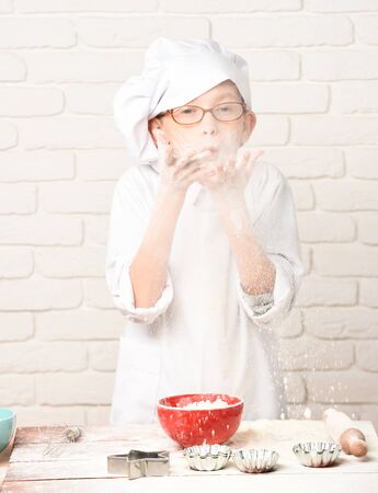 young boy small cute cook chef in white uniform and hat on happy face with glasses standing near table with rolling pin molds for cakes red bowl and playing with flour on brick wall backgroundの写真素材