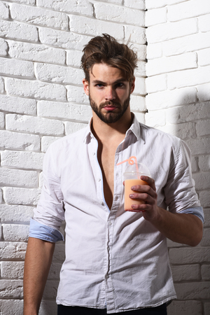young handsome bearded sexy macho man with stylish beard in white unbuttoned shirt holding glass of nonalcoholic cocktail on brick wall backgroundの写真素材