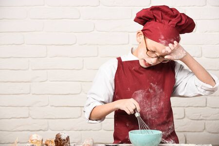 young boy cute cook chef in red uniform and hat on stained face flour with glasses standing near table with rolling pin and cooking with kitchen whisk in turquoise bowl on white brick wall backgroundの写真素材