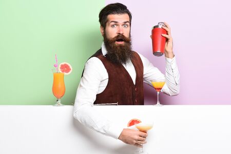 handsome bearded barman with long beard and mustache has stylish hair on surprised face holding shaker and made alcoholic cocktail in vintage suede leather waistcoat on purple green studio backgroundの写真素材