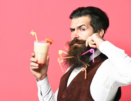 handsome bearded man with long beard and mustache has stylish hair on serious face holding glass of alcoholic beverage in vintage suede leather waistcoat on red studio backgroundの写真素材
