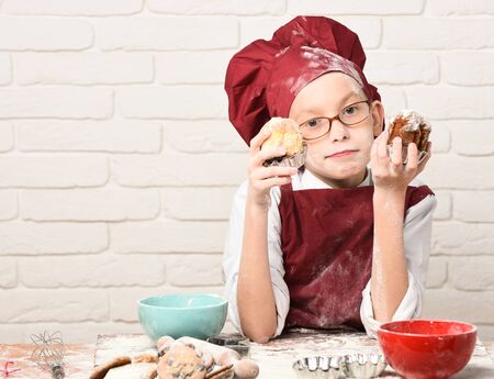 young boy cute cook chef in red uniform and hat on stained face with glasses sitting on table with colorful bowls, tasty cookies, rolling pin and holding chocolate cakes on white brick wall backgroundの写真素材