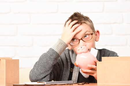 young cute pupil boy in grey sweater and glasses sitting at desk with pink piggy pig bank and...