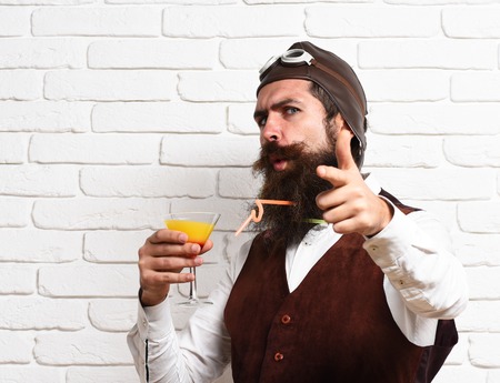 handsome bearded aviator man with long beard and mustache on funny face holding glass of alcoholic cocktail in vintage suede leather waistcoat with hat and glasses on white brick wall backgroundの写真素材