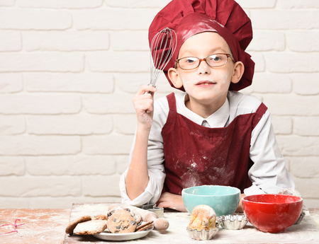 young boy cute cook chef in red uniform and hat on stained face with glasses sitting on table with colorful bowls, tasty cakes, rolling pin and kitchen whisk on white brick wall backgroundの写真素材