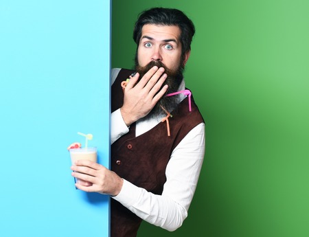 handsome bearded man with long beard and mustache has stylish hair on surprised face holding glass of alcoholic beverage in vintage suede leather waistcoat on blue green studio background, copy spaceの写真素材