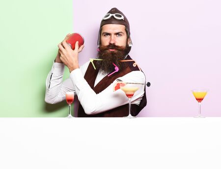 handsome bearded aviator or pilot with long beard and mustache on serious face with shaker alcoholic cocktail in vintage suede leather waistcoat with hand and glasses on purple green studio backgroundの写真素材