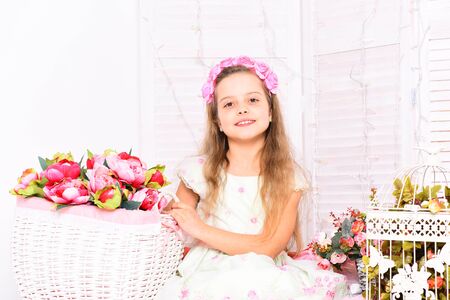 young smiling girl with wreath on curly hair and cute dress sitting with baskets of pink peonies decorative roses and cage with flowers on white brick wall background, copy spaceの写真素材