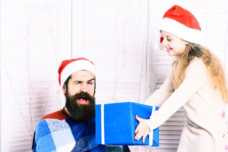 cute smiling blonde girl in beige dress gives big gift to handsome bearded man with long beard in christmas red hat in blue checkered plaid on white studio backgroundの写真素材