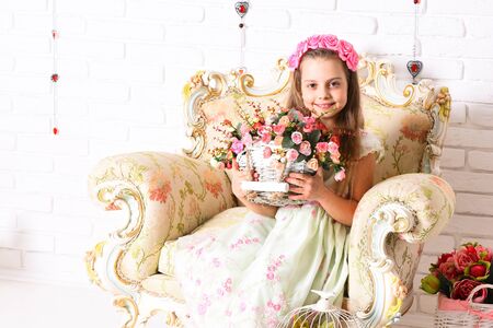 young smiling girl with wreath on curly blonde hair and cute dress sitting on vintage beige colorful armchair with basket of pink decorative roses on white brick wall backgroundの写真素材