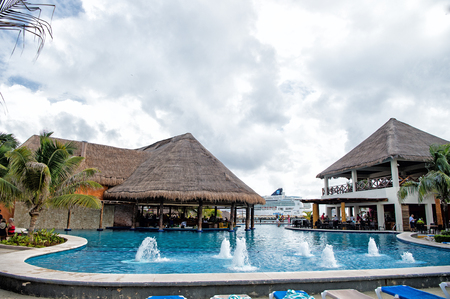 COSTA MAYA, MEXICO - December 19, 2015: swimming pool with blue water at cafe bar with straw roof with people near palm trees on cloudy sky background and cruise shipのeditorial素材
