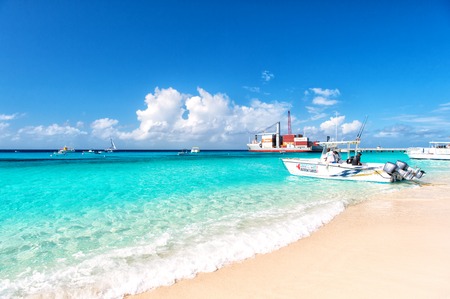 Grand Turk, Turks and Caicos Islands - December 29, 2015: big cargo ship red color with crane and many containers on ocean water and blue cloudy sky background, motor boat on sandy beach sunny outdoorのeditorial素材
