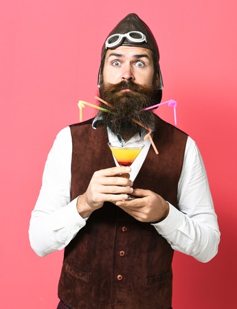 handsome bearded aviator man with long beard and mustache on surprised face holding glass of alcoholic beverage in vintage suede leather waistcoat with hat and glasses on red studio backgroundの写真素材
