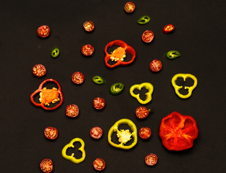 set of yellow red green fresh cut peppers rings with seeds and cherry tomatoes on black backgroundの写真素材