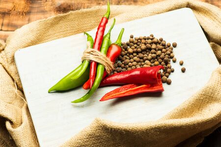 colorful fresh spicy chili peppers, black peppercorns on white paper sheet on sacking on brown...