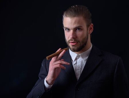 handsome bearded stylish business man in elegant black suit posing in studio with cigar on black background, copy spaceの写真素材