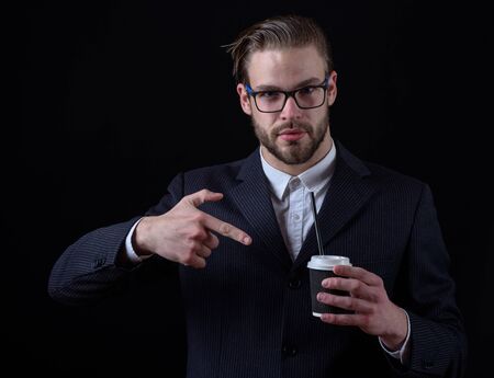 handsome bearded stylish business man in elegant black suit in glasses holding cup of coffee and shows on it in studio on black backgroundの写真素材