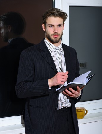 handsome young fashion bearded businessman with stylish hair in classic suit standing near window with black notebook and pen on white wall backgroundの写真素材