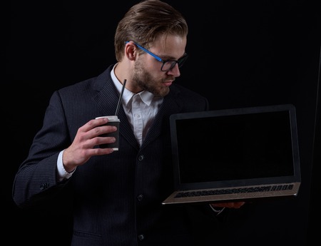 handsome bearded stylish business man in elegant black suit in glasses holding laptop with thoughtful face and coffee cup in studio on black background, copy spaceの写真素材