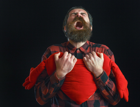 handsome bearded man or guy in checkered shirt with fashionable mustache and beard on shouting face holds valentines red heart pillow on black backgroundの写真素材