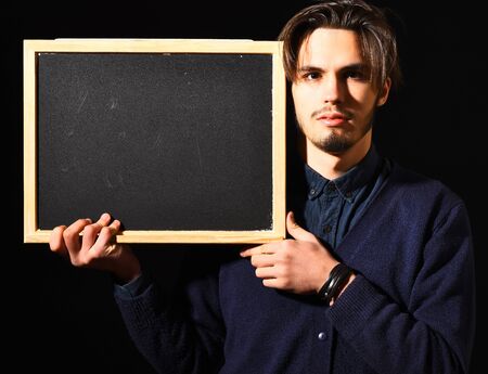 handsome bearded man in blue sweater with stylish hair on serious face holding board in black studio background, copy spaceの写真素材