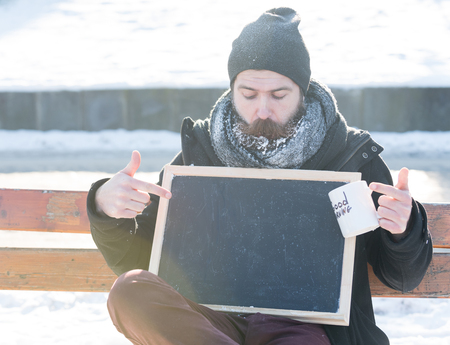 Handsome man, hipster with beard and moustache covered with white frost, points at blank black board or chalkboard holding cup with good morning text on winter day on natural background, copy spaceの写真素材