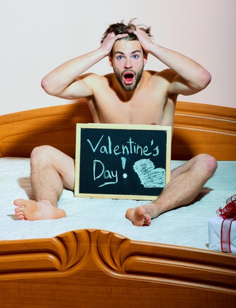 handsome bearded athletic macho man with stylish beard on surprised face sitting naked on bed with black board with inscription "valentines day" and present on pink wall background, copy spaceの写真素材