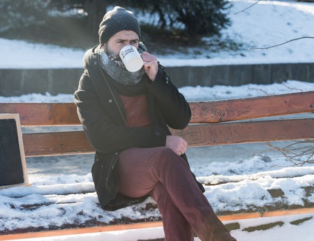 Handsome man, bearded hipster with beard and moustache covered with white frost drinks from cup with good morning text sitting on wooden bench on snowy winter day outdoors on natural backgroundの写真素材