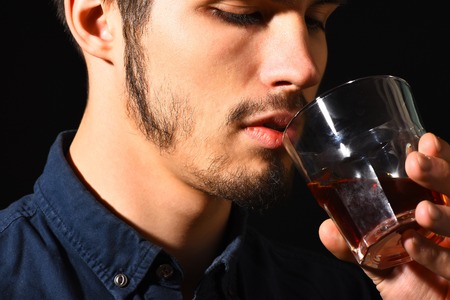 handsome bearded man in blue shirt with serious face holding and drink whisky in black studio background, close upの写真素材