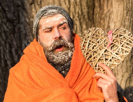 Frozen man with squinty eyes , bearded hipster, with beard and moustache covered with white frost wrapped in orange blanket with wicker heart on winter day outdoors on natural backgroundの写真素材