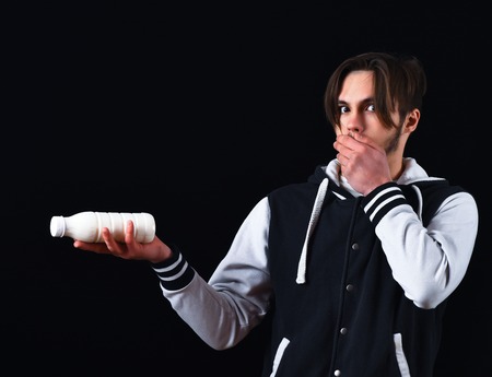 sportive handsome bearded guy in baseball jacket and surprised face holding kefir or yogurt in studio on black studio backgroundの写真素材