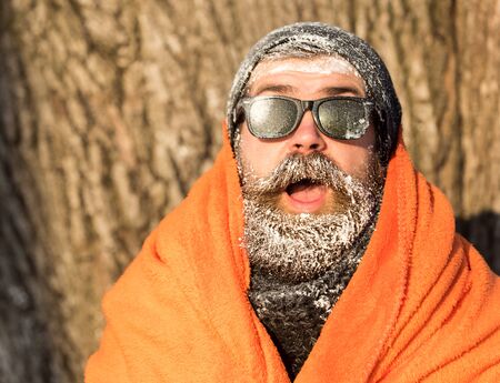 Frozen excited man, bearded hipster, with beard and moustache in black sunglasses covered with white frost wrapped in orange blanket on winter day outdoors on natural backgroundの写真素材