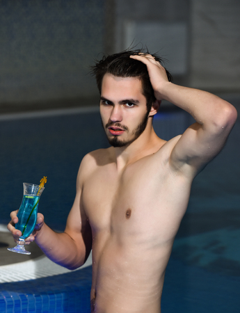 Handsome man, muscular bearded hipster or sexy macho with muscle torso drinks cocktail in glass in pool indoors on blue water backgroundの写真素材
