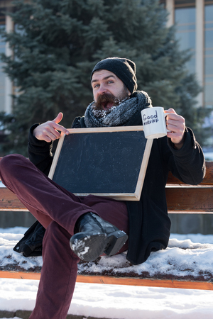 Excited man, hipster with beard and moustache covered with white frost sits on wooden bench with cup, points at blank black board or chalkboard on snowy winter day on natural background, copy spaceの写真素材