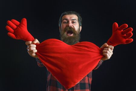 handsome bearded man or guy in checkered shirt with fashionable mustache and beard on smiling happy face holds valentines red heart pillow on black backgroundの写真素材