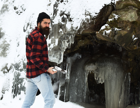 Handsome man or lumberjack, bearded hipster, with beard and moustache in red checkered shirt stands with axe at snowy stone covered with ice on winter day outdoors on natural backgroundの写真素材