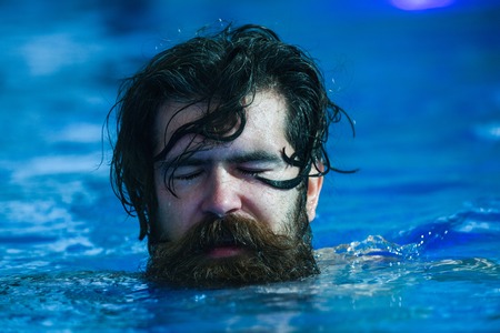 Handsome man or swimmer, bearded sexy hipster with wet beard, moustache and hair swims with closed eyes in pool indoors on blue water backgroundの写真素材