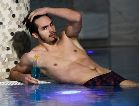 Handsome man, muscular bearded hipster or sexy macho with muscle torso, body, with cocktail in glass lies near pool indoors on blue water backgroundの写真素材