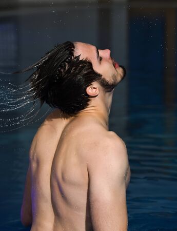 Handsome man, muscular bearded hipster or sexy macho with muscle torso splashes water from wet hair in pool indoors on blue backgroundの写真素材