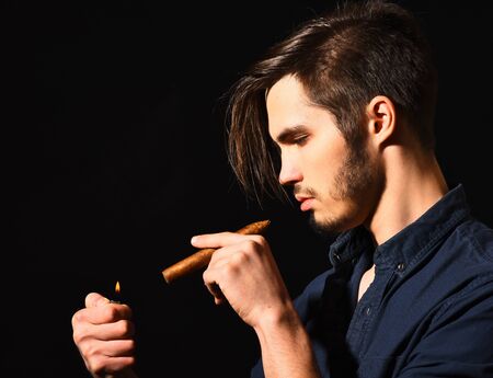handsome bearded man in blue shirt with stylish hair on serious face smoking cigar in black studio background, copy spaceの写真素材