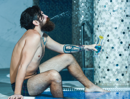 Handsome man or swimmer, bearded sexy hipster with beard and tattoo on body, bare torso, splashes cocktail from mouth in pool indoors on blue water backgroundの写真素材