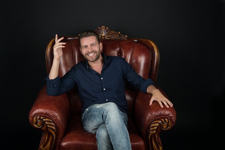 young handsome man or sexy guy with trendy beard on happy smiling face sitting on luxury brown leather chair in fashionable jeans and shirt in studio on black backgroundの写真素材
