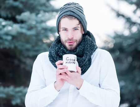 handsome bearded man or sexy guy in knitted hat, scarf and shirt in winter outdoor at green fir tree with snow holds cup with good morning text on natural backgroundの写真素材