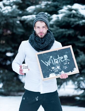 handsome bearded man or sexy guy in knitted hat, scarf and shirt in winter outdoor at green fir tree with snow holds cup with good morning text and blackboard or chalkboard on natural backgroundの写真素材