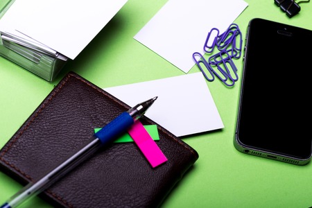 set of stationery for office: paper clips, binder clip, box with blanks, cell phone, wallet, pen, stickers on green background, side view, selective focusの写真素材