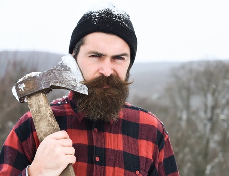 Handsome man or lumberjack, bearded hipster, with beard and moustache in red checkered shirt shaves with axe blade in snowy forest on winter day outdoors on natural backgroundの写真素材