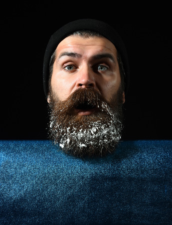 handsome bearded man or guy in winter hat with fashionable mustache on surprised face and snow in beard near blue paper on black background, copy spaceの写真素材