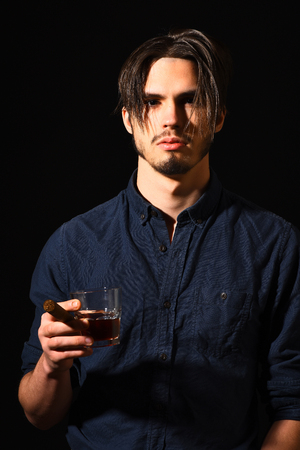 handsome bearded man in blue shirt with stylish hair on serious face holding cigar and whisky in black studio backgroundの写真素材